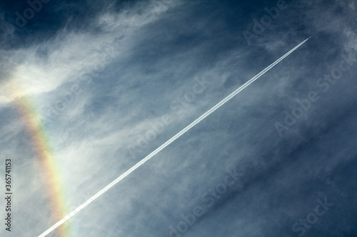 Real airplane flying overhead diagonally leaving white contrails in a blue sky with scattered clouds and a rainbow. Air traveling reopening concept.