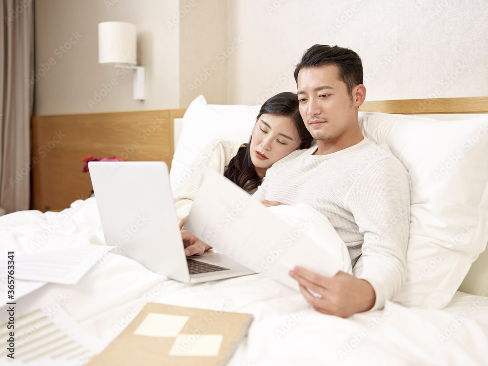 young asian businessman in bed working while wife falling asleep young asian businessman in bed working while wife falling asleep