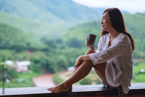 Portrait image of a beautiful asian woman holding and drinking hot coffee on balcony , looking at mountains and green nature