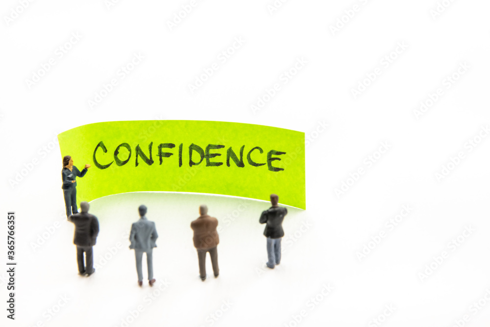Meeting with miniature figurines posed as business people standing around post-it note with Confidence handwritten message in background, minimalist abstract concept with focus on text Meeting with miniature figurines posed as business people standing around post-it note with Confidence handwritten message in background, minimalist abstract concept with focus on text