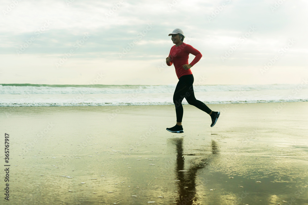 Silhouette of middle aged woman running on the beach - 40s or 50s attractive mature lady doing jogging workout enjoying fitness and healthy lifestyle at beautiful sea sunset