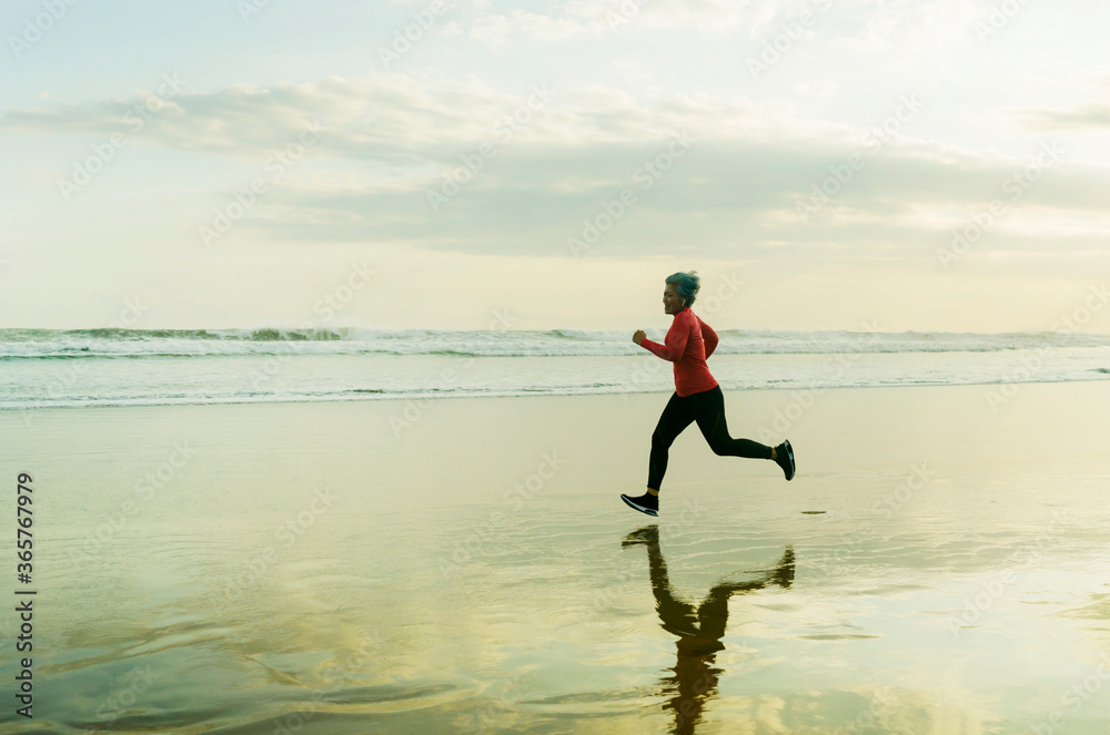 Silhouette of middle aged woman running on the beach - 40s or 50s attractive mature lady doing jogging workout enjoying fitness and healthy lifestyle at beautiful sea sunset