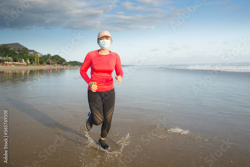 new normal running workout with face mask - attractive and happy middle aged woman on her 40s or 50s doing post quarantine jogging at beautiful beach in healthy lifestyle