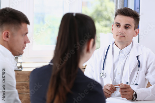 Male family doctor listen carefully young couple in office portrait. Motherhood and child delivery, new life or abortion, therapeutist reception service, physical, hospital and clinic concept