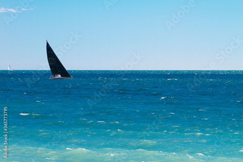A sports yacht with a black sail moving along the horizon in the azure Mediterranean Sea against a blue sky.