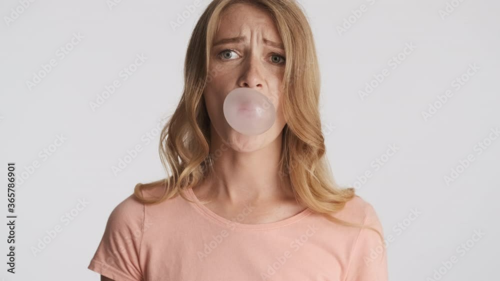 Young funny blond woman scaredly blowing bubblegum over gray background. Candy bubbles