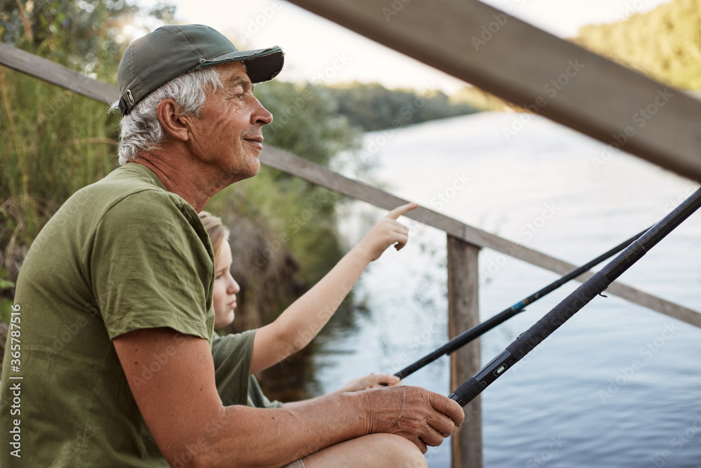 Senior man with his grandson sitting on wooden pontoon with fishing rods in hands, enjoying beautiful nature, little boy pointing at something with his finger. Senior man with his grandson sitting on wooden pontoon with fishing rods in hands, enjoying beautiful nature, little boy pointing at something with his finger.