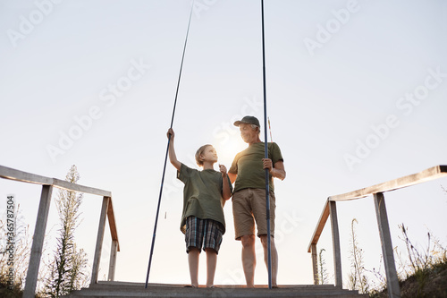 Teenage boy learning to fish with fishing rod, grandpa teaching his grandson to catch fishes, full length portrait on wooden pontoon with stairs, beautiful sunset.