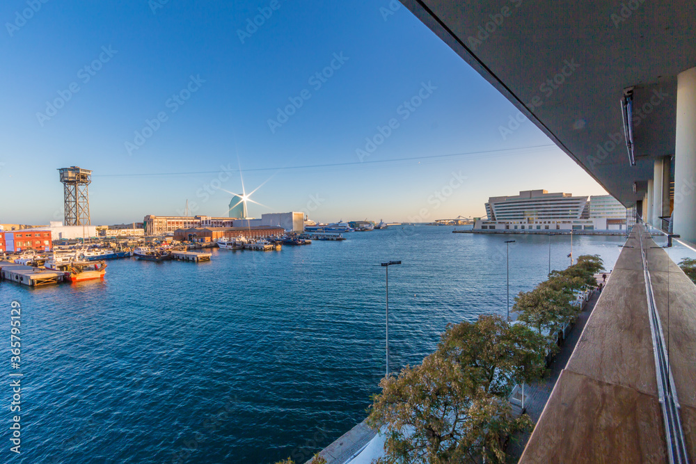 Part of the port of Barcelona with its docks, the sea seen from a building at the top, sunset with a clear blue sky with the sunlight reflected on the pier and the cable car tower in the background