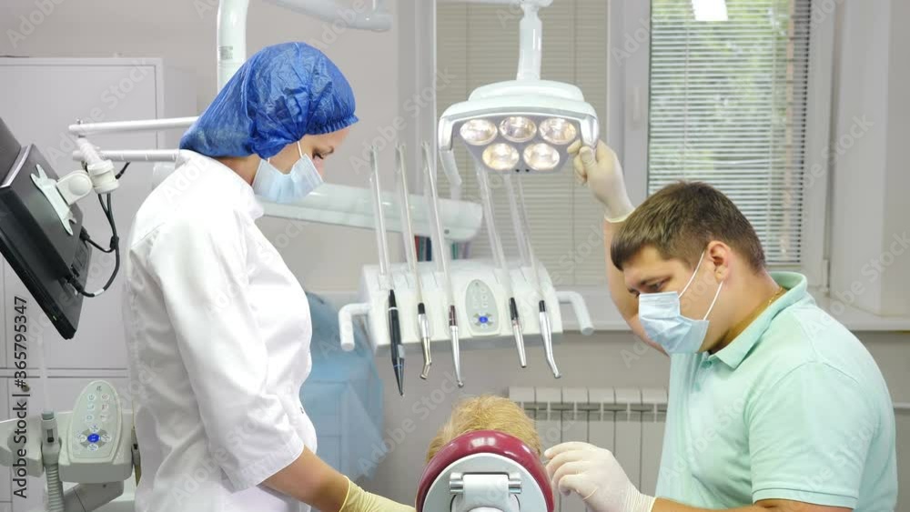 Dentistry concept. Child patient sitting on dental chair in paediatric dentists office. Assistant and male dentist working todether. Back view on Boy patient at dental clinic getting treatment. 4 k Dentistry concept. Child patient sitting on dental chair in paediatric dentists office. Assistant and male dentist working todether. Back view on Boy patient at dental clinic getting treatment. 4 k