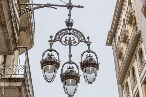 Hanging street lamp with three lamps in a black wrought iron design between buildings with balconies in a historic part of the city, gray sky on a cloudy day in Barcelona, ​​Spain