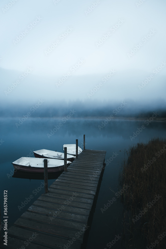 Obraz premium Boats on the lake near jetty on a misty morning. Moody scenery with boats moored in the dock with foggy forest in the background