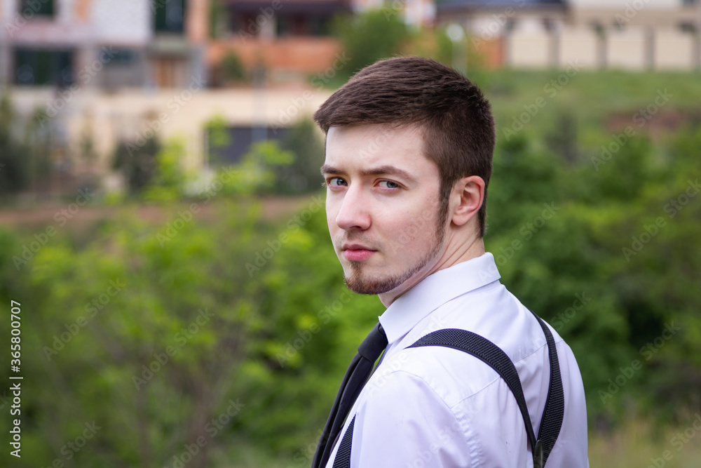Portrait of stylish brunet young man, wearing white shirt, black tie, suspenders and glasses, walking on the meadow near river. Portrait of stylish brunet young man, wearing white shirt, black tie, suspenders and glasses, walking on the meadow near river.