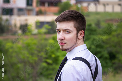 Portrait of stylish brunet young man, wearing white shirt, black tie, suspenders and glasses, walking on the meadow near river.
