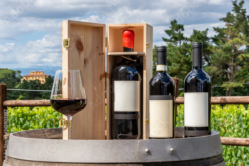Three bottles including a magnum in the wooden box and a glass of red wine on a wooden barrel with the Tuscan countryside in the background, Italy, on a sunny day photo