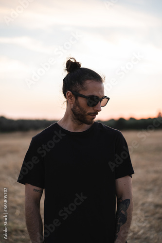 Hombre joven con gafas de sol negras, ropa negra y moño posando en un descampado seco en verano