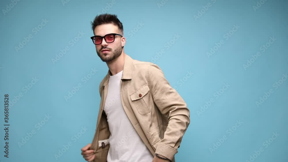 sexy young man in jacket straightening the temples and putting glasses on, adjusting jacket and holding hand behind neck in a fashion pose on blue background sexy young man in jacket straightening the temples and putting glasses on, adjusting jacket and holding hand behind neck in a fashion pose on blue background