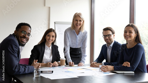Middle-aged businesswoman leader company owner and young multi ethnic professionals subordinates take break pose for camera during workshop in boardroom. Workgroup portrait. Project research concept