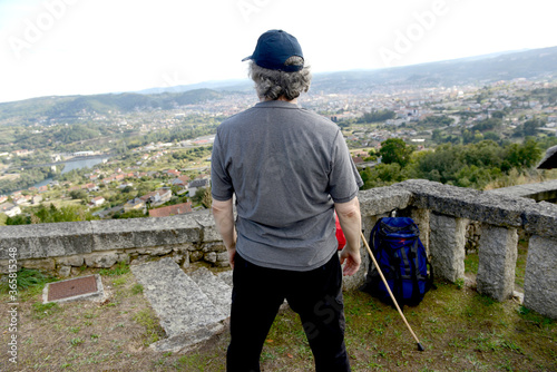 Senior man with backpack and hiking stick looks at a city from a hill photo