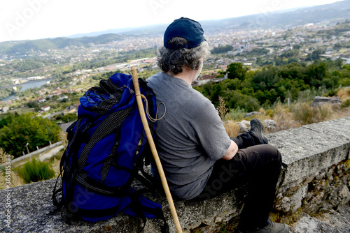 Senior man with backpack and hiking stick looks at a city from a hill photo