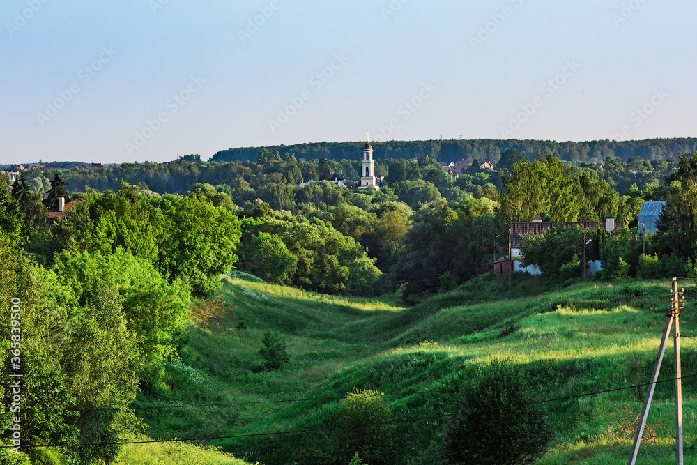 Rural landscape ravine overgrown with green grass and trees. Bell tower of Christian church in the background