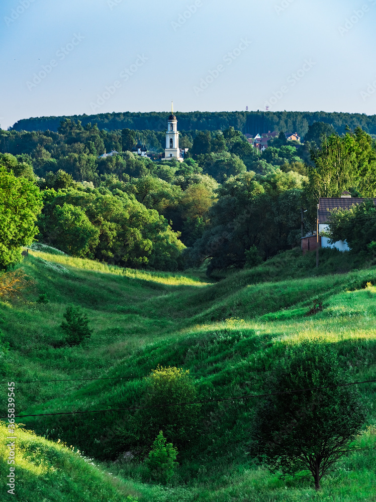 Rural landscape ravine overgrown with green grass and trees. Bell tower of Christian church in the background
