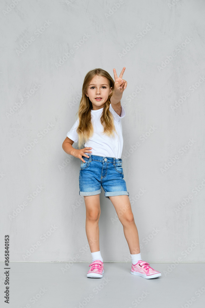 Little girl in t-shirt, jeans shorts and sneakers posing near the wall Little girl in t-shirt, jeans shorts and sneakers posing near the wall