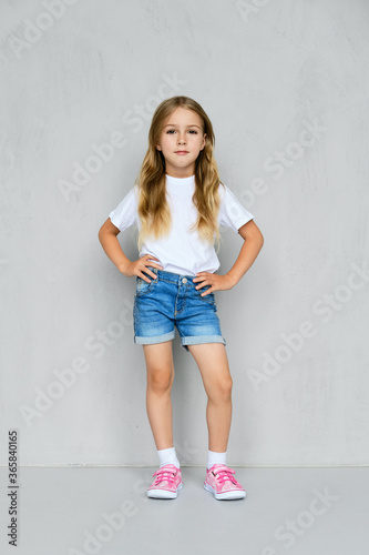 Little girl in t-shirt, jeans shorts and sneakers posing near the wall