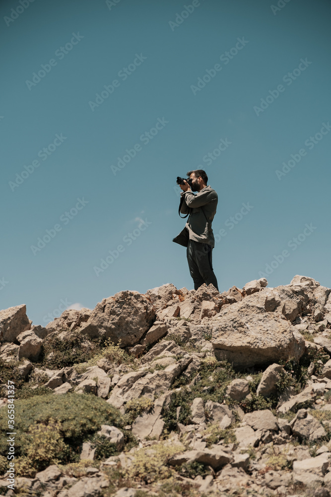 Professional photographer takes photos with camera on rocky peak with blue sky in background Professional photographer takes photos with camera on rocky peak with blue sky in background