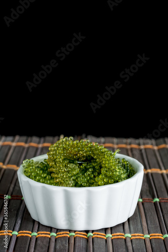 Fresh sea grapes or caviar seaweed in white bowl on wooden mat and black background. Healthy food concept.