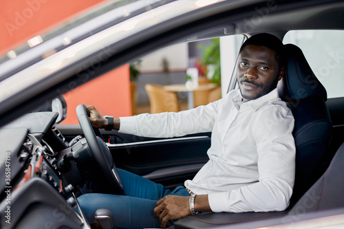 portrait of happy black businessman inside of luxurious car represented in cars showroom. man wants to buy it, likes it © alfa27