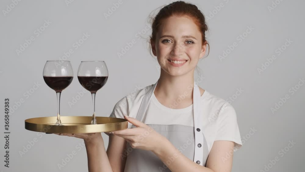 Pretty cheerful waitress in apron holding tray with wine glasses happily looking in camera isolated Pretty cheerful waitress in apron holding tray with wine glasses happily looking in camera isolated