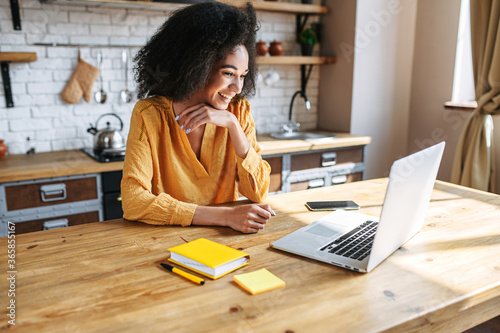 A cheerful mixed-race girl uses laptop for remote work or home leisure while sitting in the kitchen at home. Side view a nice girl with an afro hairstyle looks at screen with a smile photo