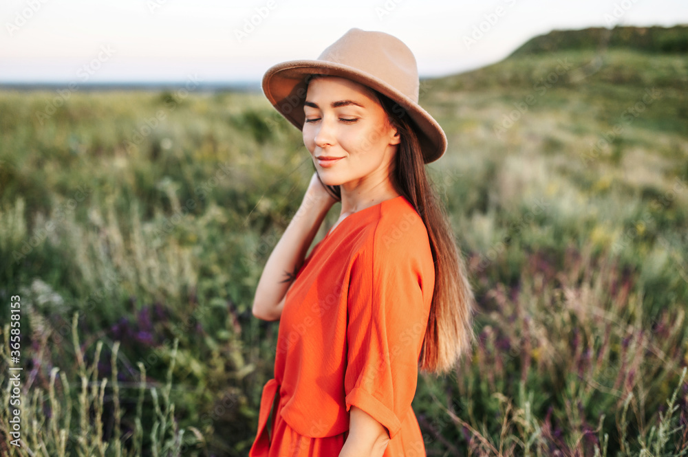 Obraz premium Romantic long haired girl in a hat is posing in the rural landscape, woman in red romper with a scenic view on the background