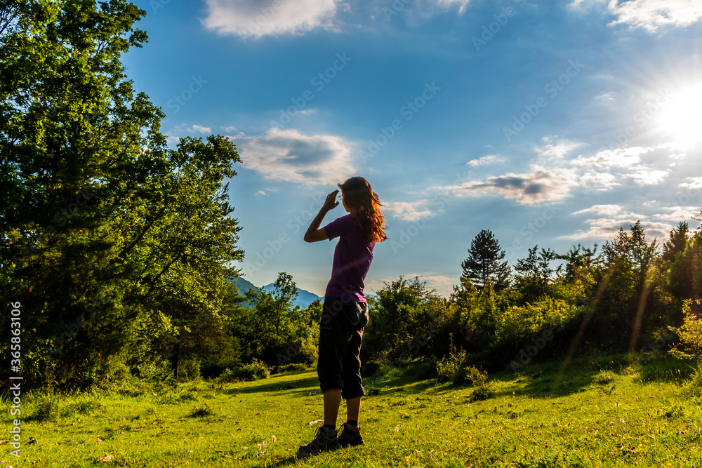 Fototapeta premium A full body shot of an unrecognizable young Caucasian redhead woman standing in a meadow in a forest in the French Alps during sunset
