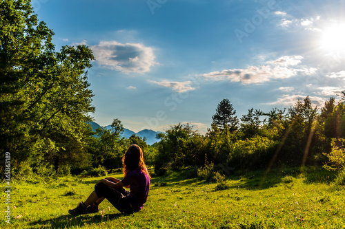 A full body shot of an unrecognizable young Caucasian redhead woman sitting on the grass in a meadow in a forest in the French Alps during sunset
