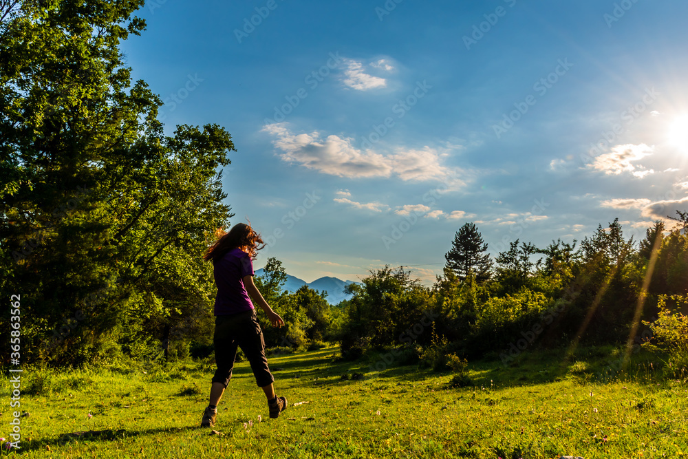 A full body shot of an unrecognizable young Caucasian redhead woman walking in a meadow in a forest in the French Alps during sunset