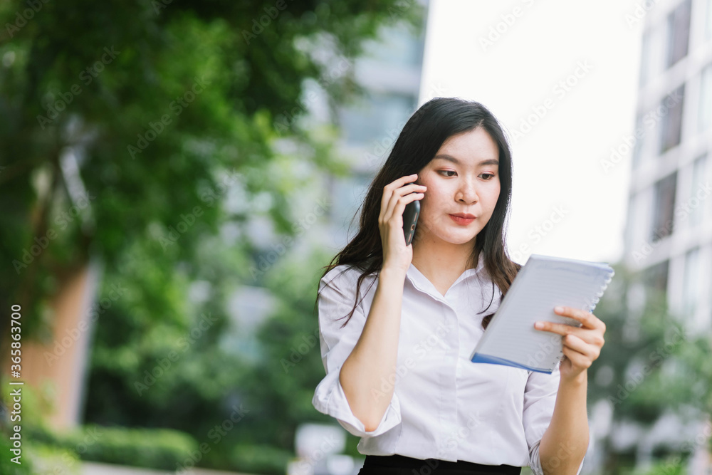 Beautiful asian business woman talking on cellphone and reading note while walking outdoor Beautiful asian business woman talking on cellphone and reading note while walking outdoor