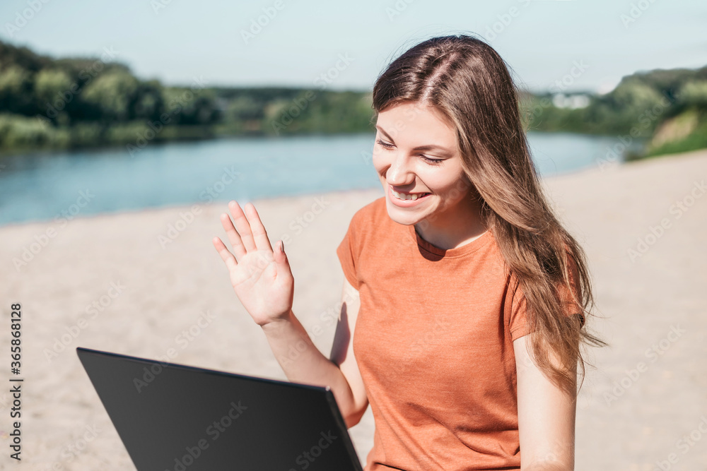 Happy young beautiful brown-haired girl in casual clothes sits on the beach with a laptop and speaks by video chat, quarantine, coronavirus