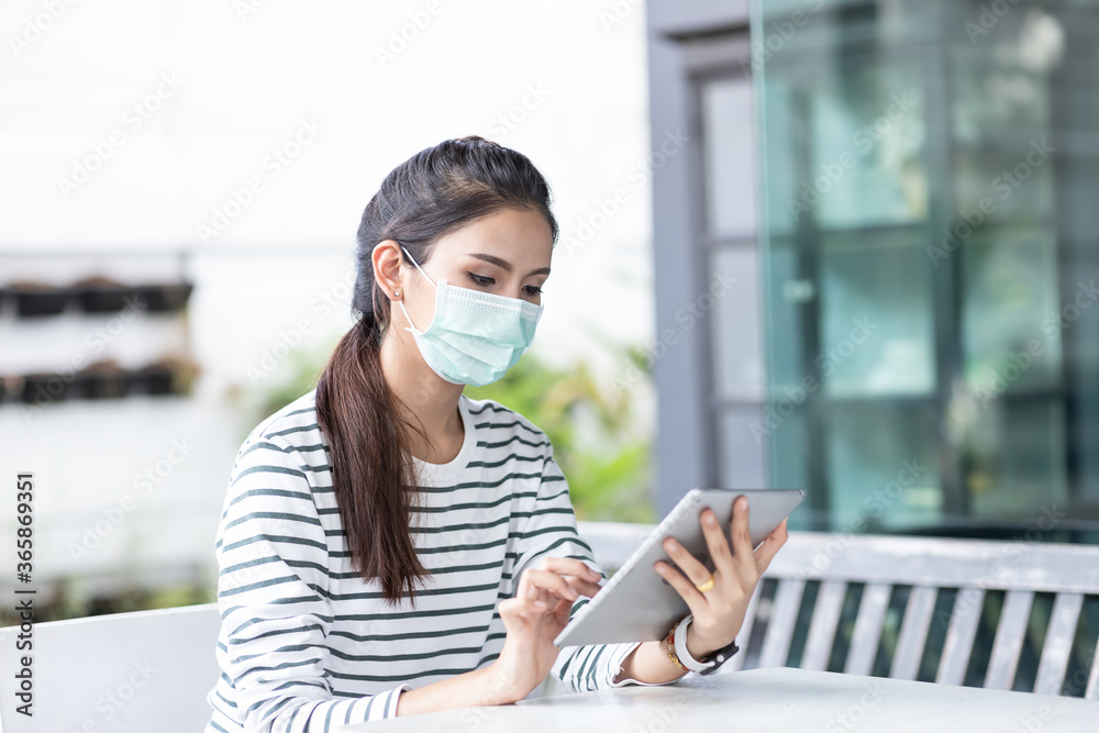 Asian young businesswoman in casual wear wearing medical face mask working with the tablet technology while sitting outside the office. Social distancing. Asian young businesswoman in casual wear wearing medical face mask working with the tablet technology while sitting outside the office. Social distancing.