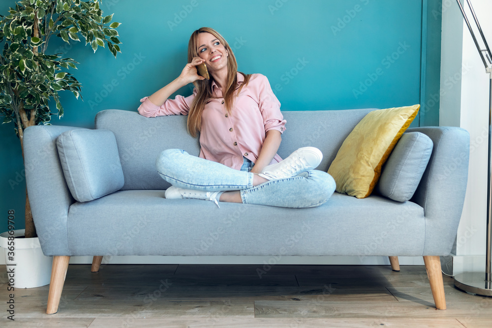 Obraz premium Smiling young woman talking on mobile phone while sitting on sofa at home.