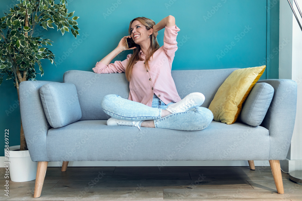 Obraz premium Smiling young woman talking on mobile phone while sitting on sofa at home.