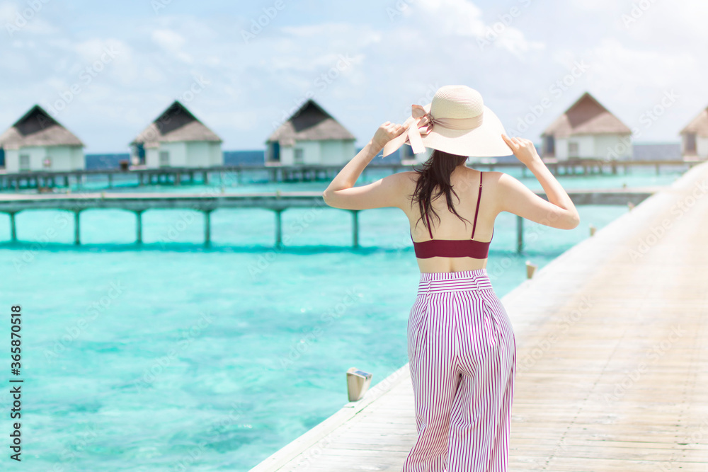 Young girl holding hat walking surrounded by beautiful turquoise ocean water. Happy traveller in tropical beach vacation. Back of young Asian woman walking over a wooden jetty in morning sunrise.
