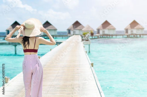 Back of a young woman walking over a wooden jetty in the morning sunrise. Young woman in hat walking surrounded by beautiful turquoise ocean water. Happy traveller woman in tropical beach vacation.