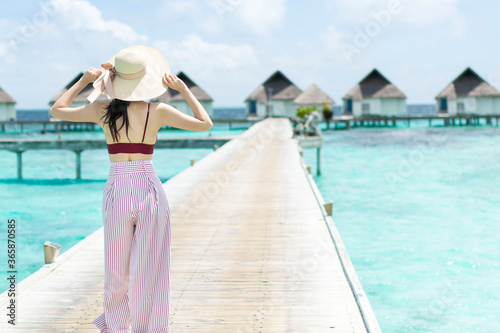 Back of a young woman walking over a wooden jetty in the morning sunrise. Young woman in hat walking surrounded by beautiful turquoise ocean water. Happy traveller woman in tropical beach vacation.
