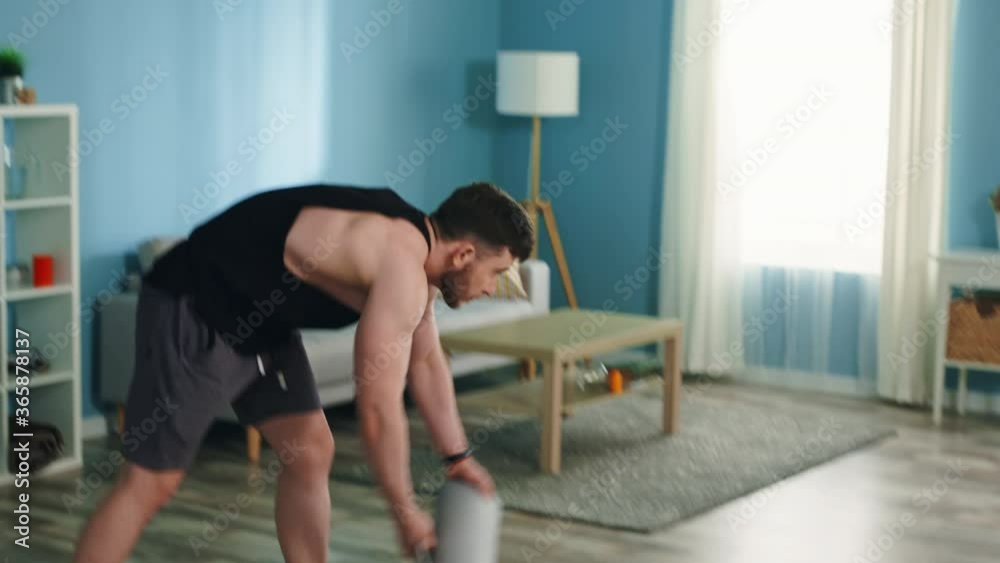 Young athletic man stays home and is going to train, using his mobile phone application, gadget, spread fitness mat, has on purpose to improve his body shape, Slow motion. Young athletic man stays home and is going to train, using his mobile phone application, gadget, spread fitness mat, has on purpose to improve his body shape, Slow motion.