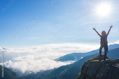 Woman with arms outstretched on mountain top, dramatic landscape clouds over the valley, clear blue sky sunburst in backlight. Summer activity fitness wellbeing freedom success.