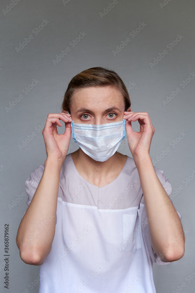 Close up of female doctor or scientist in protective facial mask over grey background. She is adjusting the medical mask with her hands Close up of female doctor or scientist in protective facial mask over grey background. She is adjusting the medical mask with her hands