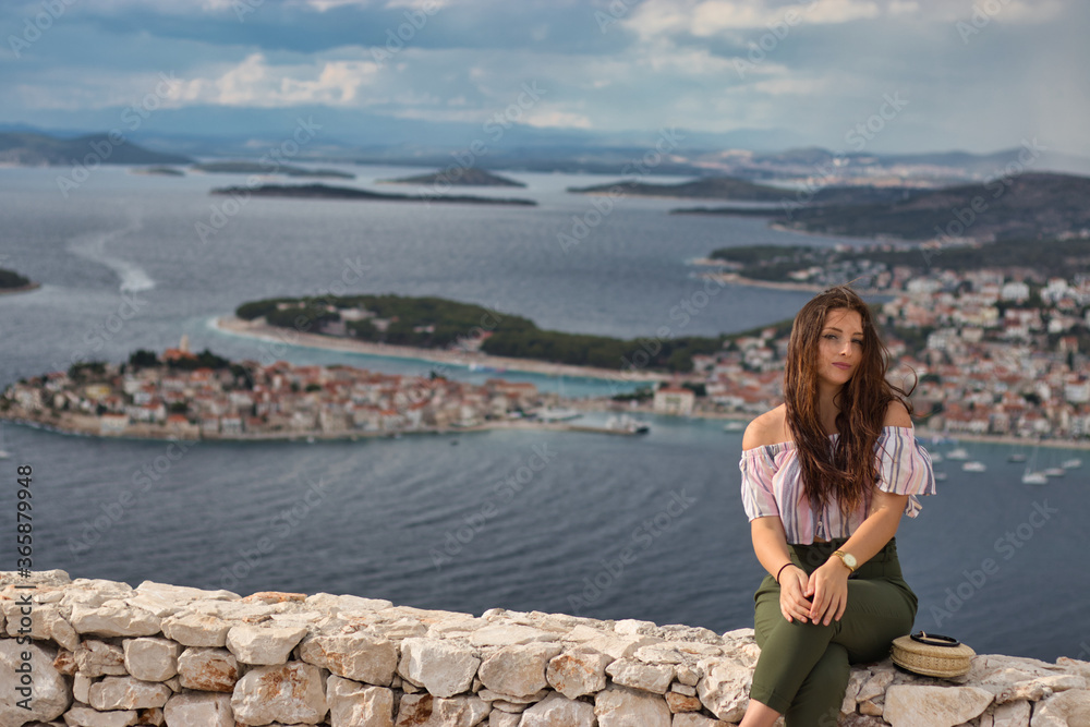 Attractive brunette sitting on a stone wall on a warm summer windy day. Fashionable girl posing with the adriatic sea and the small town of primosten in the background