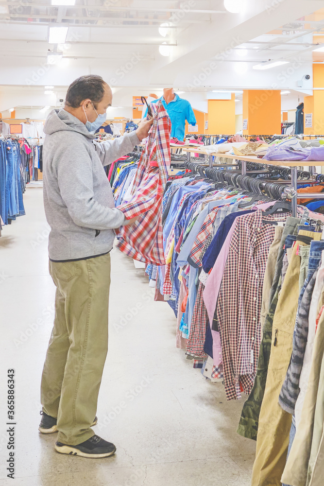 Mature man in mask choosing clothes at Second hand store.